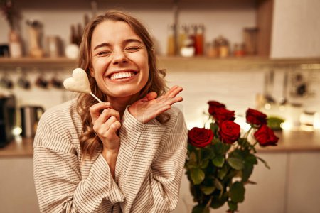 Téléchargez les photos : Joyeuse Saint-Valentin. Une jeune femme heureuse mangeant du chocolat en forme de cœur assise sur une table dans la cuisine à la maison, avec un bouquet de roses rouges debout à proximité. - en image libre de droit