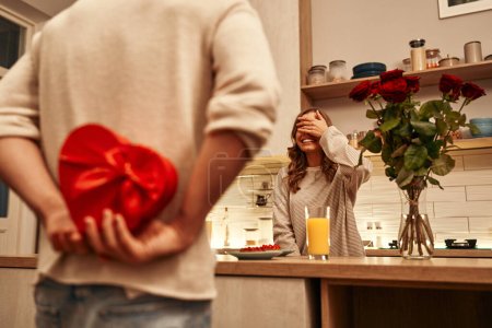 Photo for Happy Valentine's Day. A man holding a heart-shaped red gift box behind his back for his beloved woman while standing in the kitchen at home. - Royalty Free Image