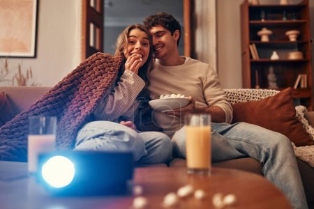 Foto de Feliz día de San Valentín. Una joven pareja enamorada sentada en el sofá de la sala de estar en casa, comiendo palomitas de maíz, cubierta con una manta y viendo una película en el proyector. - Imagen libre de derechos