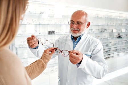 Téléchargez les photos : Médecin ophtalmologiste mature en blouse blanche démontrant des lunettes optiques à une femme dans un magasin professionnel. Correction de la vue et ophtalmologie. - en image libre de droit