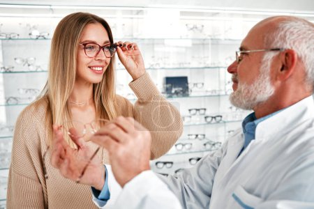 Photo for Mature ophthalmologist doctor in a white coat demonstrating optical glasses to a woman in a professional store. Vision correction and ophthalmology. - Royalty Free Image