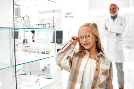 Téléchargez les photos : Une fille portant des lunettes optiques debout dans un magasin professionnel, un ophtalmologiste en arrière-plan. Correction de la vue et ophtalmologie. - en image libre de droit