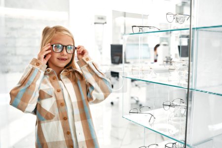 Foto de Niña linda en gafas ópticas de pie en una tienda profesional. Corrección de la visión y oftalmología. - Imagen libre de derechos