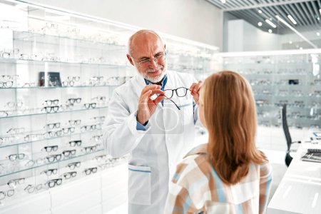 Foto de El oftalmólogo maduro demuestra gafas ópticas a una niña mientras visita una tienda profesional con varias gafas. Corrección de la vista. Atención médica, problema visual. - Imagen libre de derechos