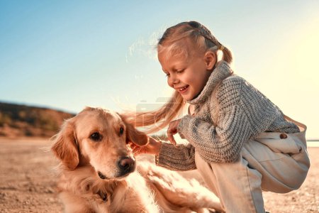 Photo for Cute little girl child having fun and playing with her golden retriever dog outside. Girl with a dog on a sandy beach. - Royalty Free Image