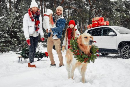 Foto de Feliz Navidad y Feliz Año Nuevo. Familia con niños y un perro de pie junto a un coche con cajas de regalos, un árbol de Navidad en un trineo, de pie sobre el telón de fondo de un bosque en clima nevado. - Imagen libre de derechos