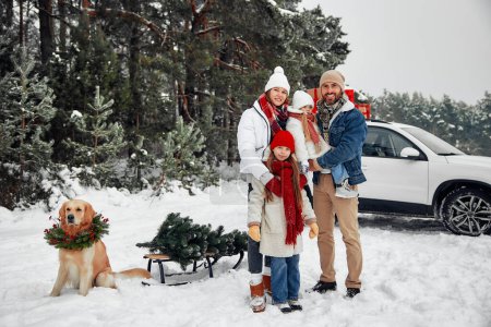 Photo for Merry Christmas and Happy New Year. Family with children and a dog standing by a car with boxes of gifts, a Christmas tree on a sleigh, standing against the backdrop of a forest in snowy weather. - Royalty Free Image