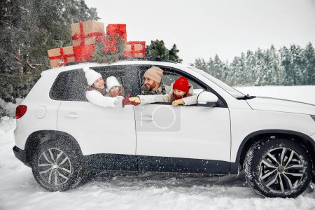 Foto de Feliz Navidad y Feliz Año Nuevo. Familia feliz con niños sentados en un coche cargado de cajas de regalos y un árbol de Navidad sobre el fondo de un bosque con clima nevado. - Imagen libre de derechos