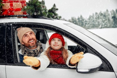 Téléchargez les photos : Joyeux Noël et bonne année. Joyeux famille avec des enfants assis dans une voiture chargée de boîtes de cadeaux et un arbre de Noël sur le fond d'une forêt par temps neigeux. - en image libre de droit