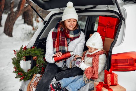 Téléchargez les photos : Joyeux Noël et bonne année. Maman et fille sont assis sur une couverture chaude dans le coffre ouvert d'une voiture avec un tas de cadeaux autour et boire du thé d'un thermos par temps neigeux. - en image libre de droit