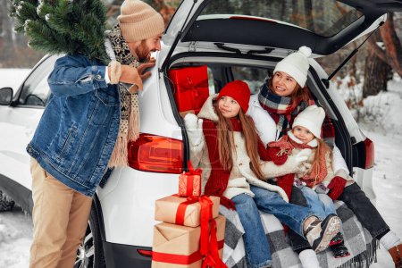 Photo for Merry Christmas and Happy New Year. A family with children sitting on a warm blanket in the open trunk of a car with a bunch of gifts around, a man holding a Christmas tree in snowy weather. - Royalty Free Image