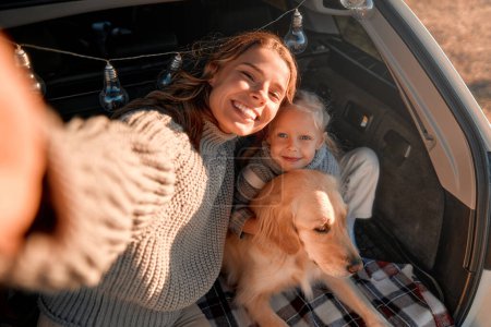 Foto de Una joven madre con su linda hija y perro recuperador sentada en una manta caliente en el maletero abierto de un coche y tomando una selfie en el teléfono. Familia viajando con una mascota. - Imagen libre de derechos