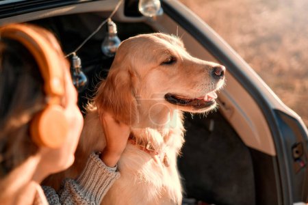Foto de Mujer feliz sentada en el maletero abierto de un coche con su perro en una manta caliente, escuchando música en los auriculares. Mujer viajando con mascota. - Imagen libre de derechos