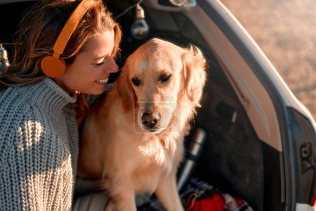 Téléchargez les photos : Une femme heureuse assise dans le coffre ouvert d'une voiture avec son chien sur une couverture chaude, écoutant de la musique sur un casque. Femme voyageant avec animal de compagnie. - en image libre de droit