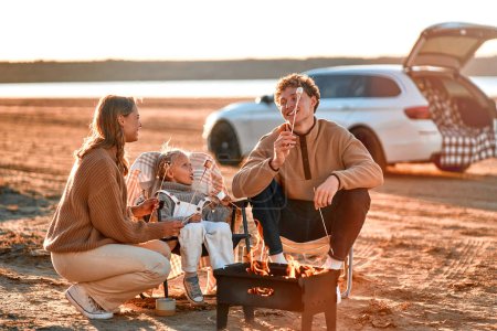 Photo for A family with a daughter sitting at the grill and roasting marshmallows. In the background is a car with an open trunk. Family traveling. Travel, vacation and tourism concept. - Royalty Free Image