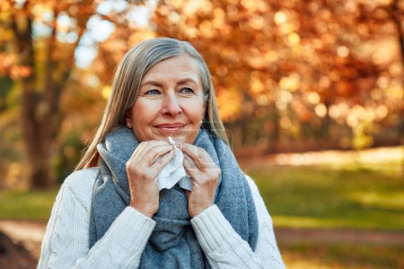 Foto de Una mujer madura de pelo gris cayó enferma, limpiándose la nariz con un pañuelo en un parque en otoño dorado. Goteo nasal y frío. - Imagen libre de derechos