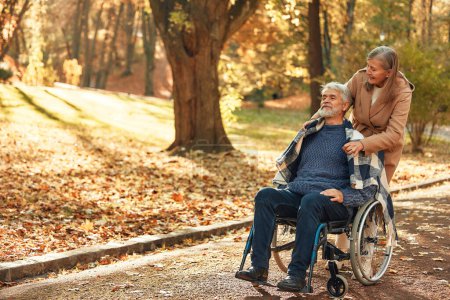 Téléchargez les photos : Belle femme âgée marchant avec son mari dans un fauteuil roulant dans le parc en automne. Aide et soins en réadaptation. - en image libre de droit