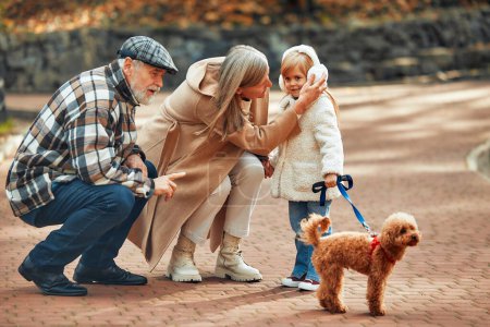 Foto de Pareja madura paseando con un niño y un perro en el parque. Abuelos paseando con su nieta y su perro en el parque en otoño. - Imagen libre de derechos
