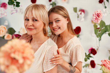Photo for Happy Mother's Day and Women's Day. Mom and daughter in dresses standing hugging isolated on a white background, there are many flowers in the air around. - Royalty Free Image