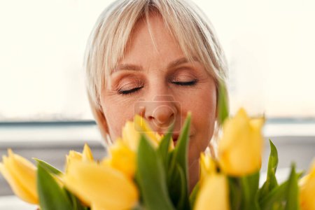 Photo for Happy Mother's Day and Women's Day. Charming mature woman sitting with a bouquet of tulips in the living room by the window. Mother's love and care. - Royalty Free Image