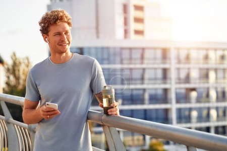 Téléchargez les photos : Un homme boit de l'eau et utilise un smartphone tout en se reposant après avoir couru à l'extérieur sur un pont. Exercices matinaux et jogging. Sports et loisirs, mode de vie actif. - en image libre de droit