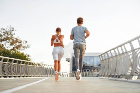 Téléchargez les photos : Un couple en tenue de sport faisant du jogging à l'extérieur sur un pont. Exercices matinaux et jogging. Sports et loisirs, mode de vie actif. - en image libre de droit