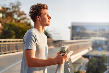 Téléchargez les photos : Un homme boit de l'eau et utilise un smartphone tout en se reposant après avoir couru à l'extérieur sur un pont. Exercices matinaux et jogging. Sports et loisirs, mode de vie actif. - en image libre de droit