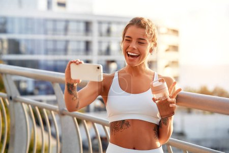 Téléchargez les photos : Une femme avec une bouteille d'eau prenant un selfie sur un smartphone tout en se reposant après avoir couru à l'extérieur sur un pont. Exercices matinaux et jogging. Sports et loisirs, mode de vie actif. - en image libre de droit