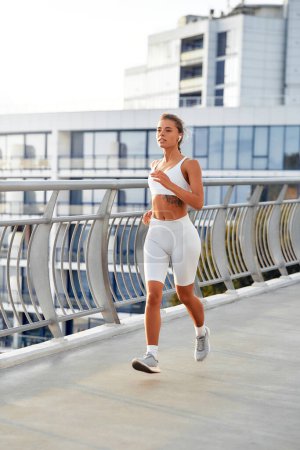 Téléchargez les photos : Femme en tenue de sport avec écouteur courant sur un pont. Exercices matinaux et jogging. Sports et loisirs, mode de vie actif. - en image libre de droit