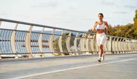 Téléchargez les photos : Une femme en tenue de sport avec un écouteur courant sur un pont au bord de la mer. Exercices matinaux et jogging. Sports et loisirs, mode de vie actif. - en image libre de droit