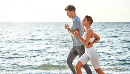 Téléchargez les photos : Un couple en vêtements de sport courant le long du rivage par la mer. Exercices matinaux et jogging. Sports et loisirs, mode de vie actif. - en image libre de droit