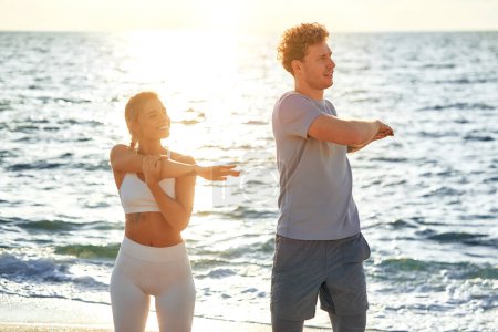 Téléchargez les photos : Couple en tenue de sport faisant des exercices d'échauffement sur le rivage par la mer. Exercices matinaux et jogging. Sports et loisirs, mode de vie actif. - en image libre de droit