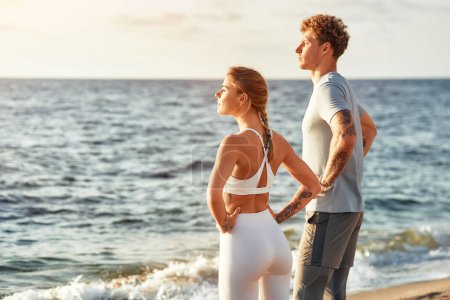 Téléchargez les photos : Couple en tenue de sport faisant des exercices d'échauffement sur le rivage par la mer. Exercices matinaux et jogging. Sports et loisirs, mode de vie actif. - en image libre de droit
