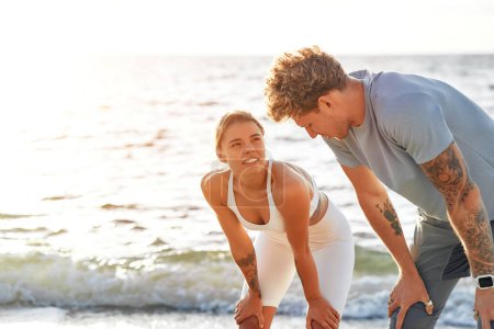 Téléchargez les photos : Un couple en tenue de sport se reposant après avoir couru sur le rivage par la mer. Exercices matinaux et jogging. Sports et loisirs, mode de vie actif. - en image libre de droit