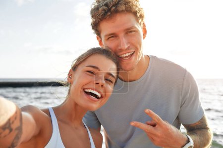Photo for A couple in sportswear relaxing after running on the shore by the sea and taking a selfie on a smartphone. Morning exercises and jogging. Sports and recreation, active lifestyle. - Royalty Free Image