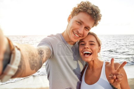Téléchargez les photos : Un couple en tenue de sport se détendre après avoir couru sur le rivage par la mer et prendre un selfie sur un smartphone. Exercices matinaux et jogging. Sports et loisirs, mode de vie actif. - en image libre de droit