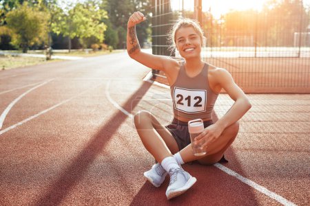 Photo for A woman in sportswear with a number resting after a run at the stadium. Morning exercises and jogging. Sports and Olympic Games, active lifestyle. - Royalty Free Image