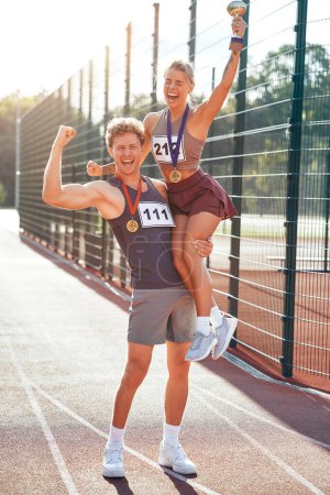 Photo for A couple of winners in sportswear with numbers and gold medals for first place and a cup at the stadium celebrating after the competition. Sports and Olympic Games, victory concept. - Royalty Free Image