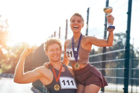 Photo for A couple of winners in sportswear with numbers and gold medals for first place and a cup at the stadium celebrating after the competition. Sports and Olympic Games, victory concept. - Royalty Free Image