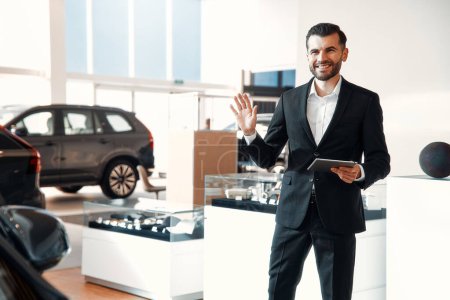 Photo for A salesman in a suit with a tablet standing in a car showroom waving his hand to greet customers and smiling. Buying, renting and insuring a car. - Royalty Free Image