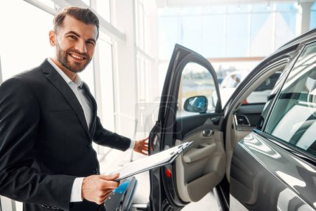 Photo for A salesman in a suit with documents stands in a car showroom and demonstrates the car to buyers, opening the doors. Buying, renting and insuring a car. - Royalty Free Image