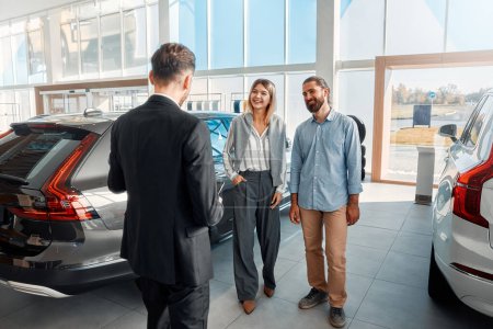 Photo for A salesman in a suit with a tablet consulting a couple of clients in a car dealership. Purchase, rental and insurance of a car. - Royalty Free Image