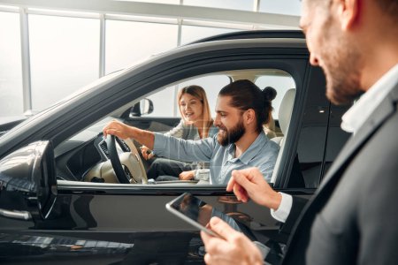 Photo for A young couple is sitting inside a car and examining it in a car showroom and receiving advice from a salesperson with a tablet. Purchase, rental and insurance of a car. - Royalty Free Image