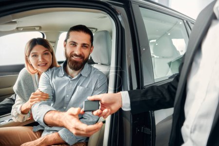 Photo for Happy young couple sitting in a new car at a car dealership and receiving the keys from the seller. Purchase, rental and insurance of a car. - Royalty Free Image