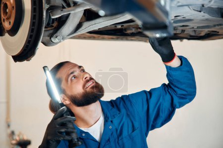 Photo for View from side of concentrated worker of car service checking running gear of automobile. Male mechanic in uniform and gloves standing under car, keeping lamp and looking for breakage. - Royalty Free Image