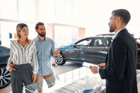 Photo for A young couple entered a car dealership to buy a new car. A salesman in a suit advising a couple and helping them choose a car. Buying, renting a car. - Royalty Free Image