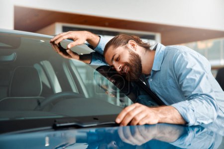 Photo for Happy young man hugging his new car in showroom. Satisfied guy with closed eyes embracing the hood of the automobile. Dreaming man lying on car bonnet hugging it. - Royalty Free Image