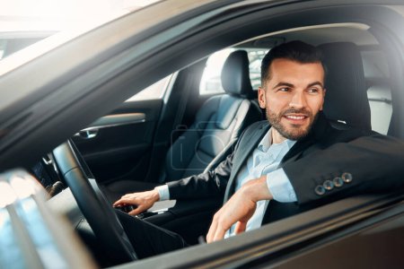 Téléchargez les photos : Un bel homme barbu homme d'affaires en costume assis au volant d'une voiture et souriant. - en image libre de droit