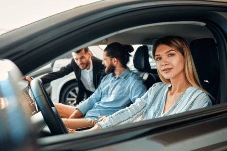 Foto de Una pareja está sentada en un coche en un concesionario de automóviles y recibiendo consejos de un vendedor. Compra, alquiler y seguro de un coche. - Imagen libre de derechos