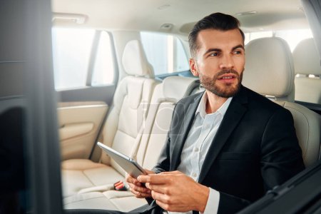 Téléchargez les photos : Un homme d'affaires en costume noir assis dans une voiture de luxe avec un intérieur en cuir blanc et utilisant une tablette. - en image libre de droit